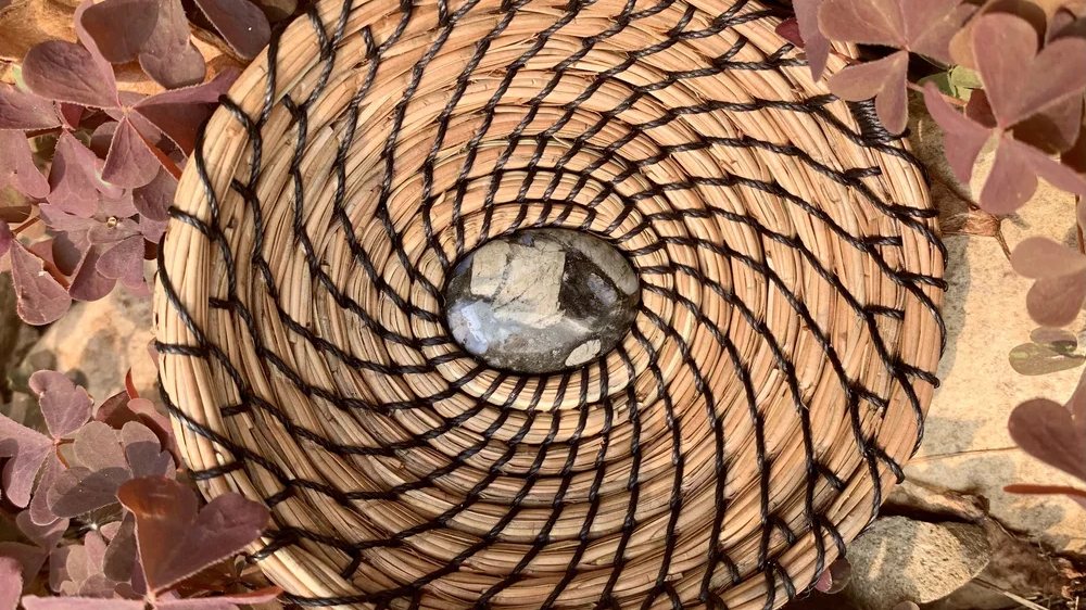 A round coiled basket of pale pine needles stitched in a black spiral, a polished grey stone set at its centre, resting among deep red clover leaves.