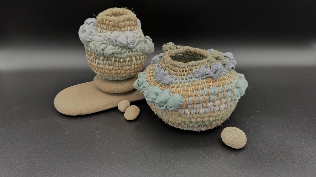Two small coiled baskets made from pale sage, blue-grey and sand-coloured fabric strips, their rims ruffled with looped cloth, resting on smooth stones against a dark background.