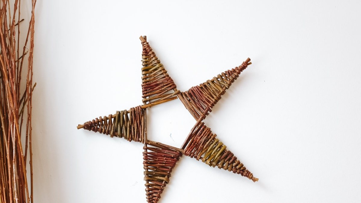 A woven willow five-pointed star hanging on a pale wall beside a bundle of willow rods.