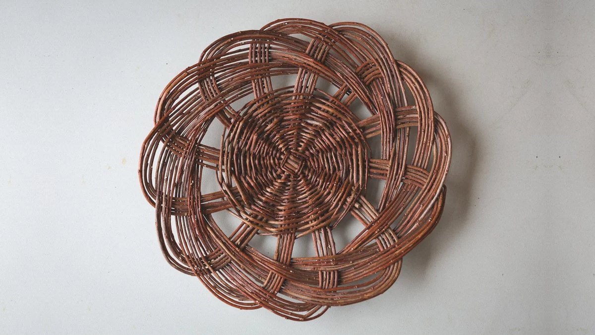 A round willow tray woven in a scalloped flower pattern, seen from above against a pale wall.