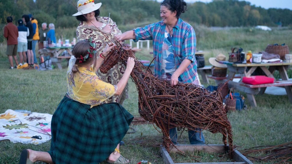 Two weavers lifting the head onto a large willow goat sculpture on the grass at the harvest festival, picnic tables and visitors behind them.