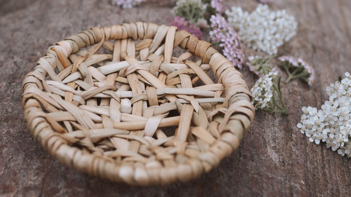 A shallow round basket woven from flat pale rush, with a coiled rim and an open lattice base, resting on weathered wood beside sprigs of white and pink yarrow.